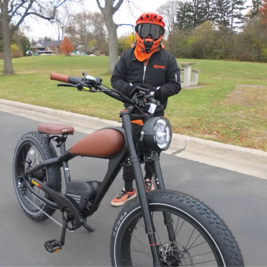 Person wearing an orange helmet and black jacket with a black electric bike on a road. Cheetah Pro Sale Revi Ebike