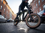 A person riding an electric fat-tire bicycle on a cobblestone street between brick apartment buildings. The bike has a matte olive green frame, black components, and tan sidewall tires. Revi Cougar 
