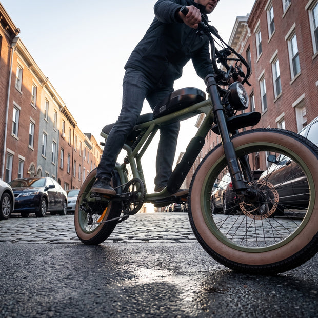 A person riding an electric fat-tire bicycle on a cobblestone street between brick apartment buildings. The bike has a matte olive green frame, black components, and tan sidewall tires. Revi Cougar 