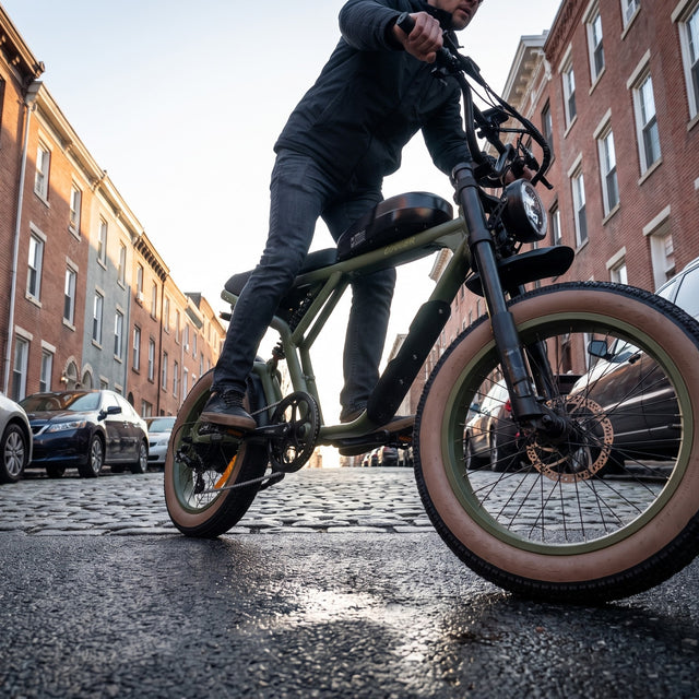 A person riding an electric fat-tire bicycle on a cobblestone street between brick apartment buildings. The bike has a matte olive green frame, black components, and tan sidewall tires. Revi Cougar 