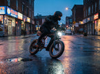 A person riding an electric bicycle on a wet city street at dusk, wearing a black helmet and dark jacket. The bike has a sleek black frame and a bright front headlight, with storefront lights reflecting on the rain-slicked road. The new Revi Cougar e-Wake 
