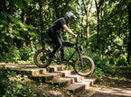 A person riding a black electric mountain bike with fat tires down a set of stone steps in a dense green forest. Back to modern Revi e-bikes 