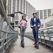 Two commuters riding electric scooters with ROCKBROS hard shell bags mounted on the stems
